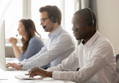 Serious african call center agent in wireless headset working on computer with diverse team, focused black male operator telemarketer using pc in customer support service group office, helpdesk